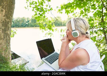 Eine ältere Geschäftsfrau, die im Sommer am Seeufer mit einem Laptop unterwegs war, während sie nachdachte Stockfoto