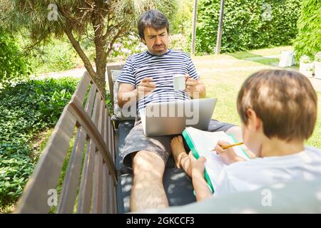 Freier Mitarbeiter als Vater mit Computer und Sohn auf einer Bank im Sommer im grünen Garten Stockfoto