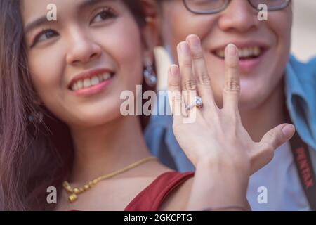 Glückliche junge Frau, die am Finger Diamantringe an Freund Selfie am Telefon zeigt. Neu verlobt Mädchen und Verlobten im Hintergrund. Verlobte Liebe und ein Stockfoto