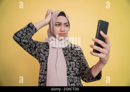 Junge Frau im Hijab verwirrt, während sie auf den Smartphone-Bildschirm mit kratzender Hand schaute Stockfoto