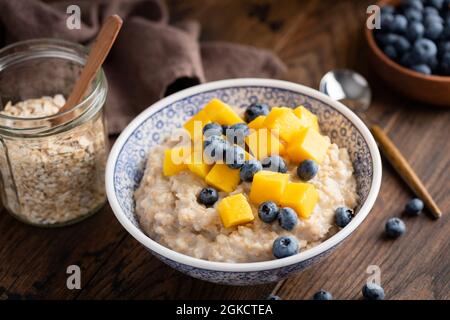 Gekochter Haferbrei mit Heidelbeeren und Mango in einer Schüssel auf einem dunkelbraunen Holztisch im Hintergrund, Nahaufnahme Stockfoto