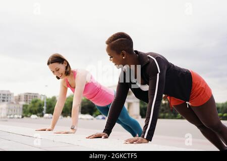 Zwei lächelnde, vielfältige junge Frau in Athletic Workout Clothes machen eine Plank-Übung, die gemeinsam auf dem Stadtplatz trainiert. Stockfoto