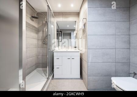 WC in moderner Wohnung mit Dusche, Bildschirm, weißem Schrank, WC und Wänden mit grauen Fliesen bedeckt Stockfoto