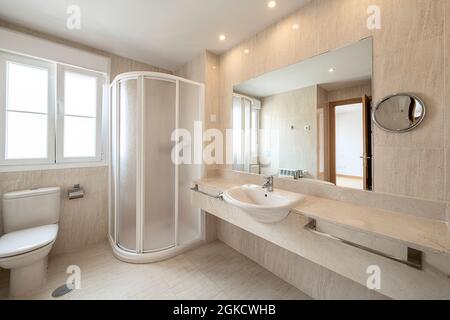 Badezimmer mit Dusche mit weißem Bildschirm in der Ecke, cremefarbenem Marmor an den Wänden und am Boden und Arbeitsplatte in der gleichen Farbe. Stockfoto