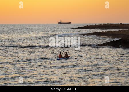 Zwei Mädchen sitzen auf dem Brett im ruhigen mittelmeer bei Sonnenuntergang in der Stadt paphos auf zypern. Silhouetten von 2 Mädchen, die auf dem Paddelbrett paddeln Stockfoto