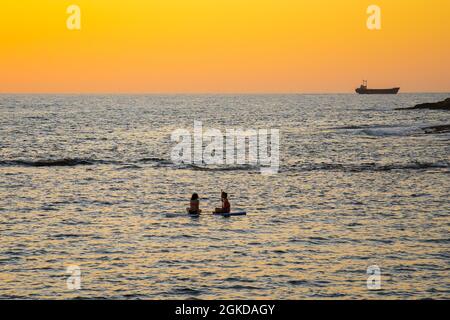 Zwei Mädchen sitzen auf dem Brett im ruhigen mittelmeer bei Sonnenuntergang in der Stadt paphos auf zypern. Silhouetten von 2 Mädchen, die auf dem Paddelbrett paddeln Stockfoto