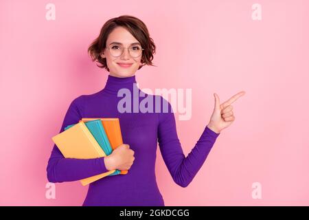 Foto von niedlichen Brünette Millennial Dame halten Bücher Punkt leeren Raum tragen Brille violetten Pullover auf rosa Hintergrund isoliert Stockfoto