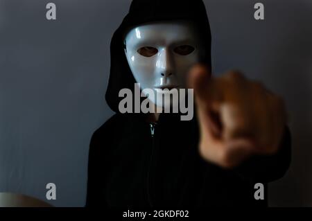 Mann mit einer schwarzen Kapuzenjacke, die eine weiße Halloween-Maske trägt und mit einem Zeigefinger auf dunklem Hintergrund Stockfoto