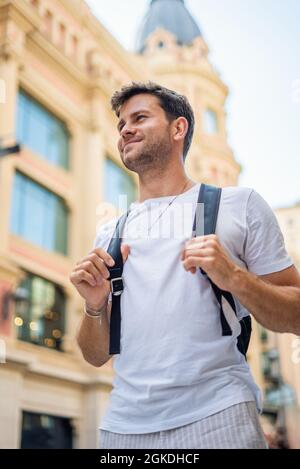 Ein kleiner, fröhlicher junger bärtiger männlicher Reisender in weißem T-Shirt mit Rucksack genießt den Sommerurlaub in der Stadt, während er urbane Straßen mit alter Architektur erkundet Stockfoto