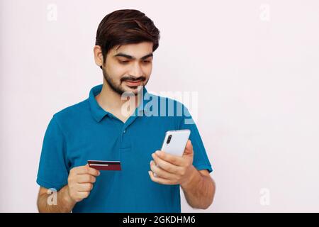 Ein gutaussehender Mann bezahlt mit Kreditkarte und Smartphone. Foto eines indischen Mannes im T-Shirt auf isoliertem Hintergrund. Stockfoto