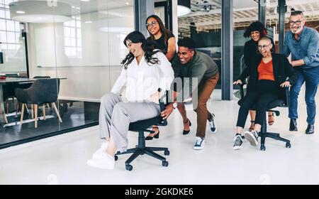 Geschäftsleute, die im Büro Spaß haben. Fröhliche Gruppe von Kollegen, die sich an einem modernen Arbeitsplatz auf Stühlen schieben. Geschäftsteam Stockfoto