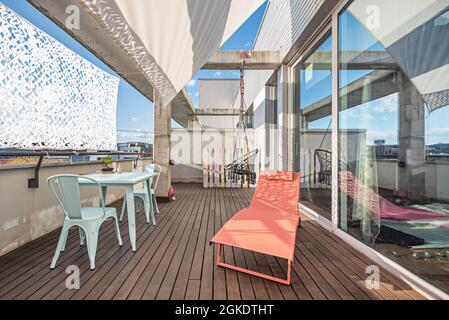 Terrasse en un Loft Penthouse mit großem Fenster, Glastür, Gartentisch und Stühle, orange Hängematte und graue Schaukel. Stockfoto