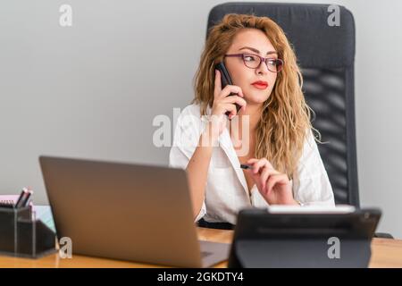 Die junge, schöne Geschäftsfrau spricht mit dem Telefon und sitzt selbstbewusst auf ihrem Schreibtisch, um ihre Arbeit zu überprüfen. Hochwertige Fotos Stockfoto