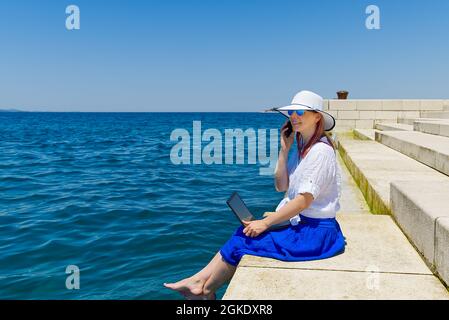 Arbeiten Sie von überall aus. Seitenansicht einer jungen Frau, Freiberuflerin im Strohhut, die am Laptop arbeitet, während sie am Strand sitzt Stockfoto
