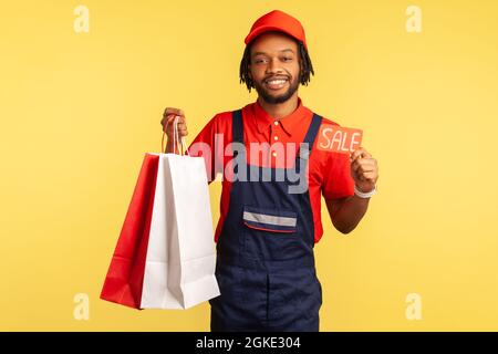 Lächelnder Kurier in Uniform hält Einkaufstaschen aus dem Modeladen und zeigt Verkaufscoupon, Verkäufe bei Online-Bestellungen und Lieferung. Innenstudio s Stockfoto