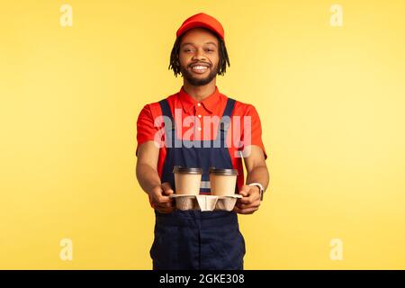 Lieferservice. Freundlicher, fröhlicher Kurier in blauen Overalls, der Kaffee anbietet, Getränke in Einwegbechern gibt und schnell leckere Getränke liefert. Zoll Stockfoto