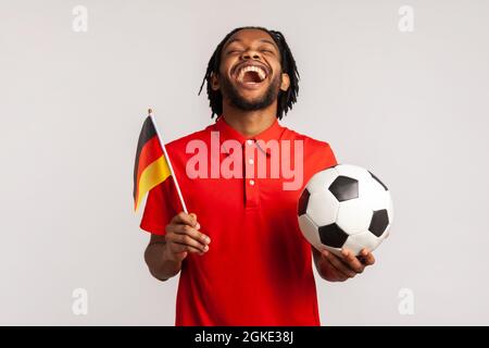 Schreiender Mann in einem roten lässigen T-Shirt, Fußballfan freut sich aufrichtig, unterstützt die Lieblingsmannschaft mit Ball und deutscher Flagge, beim Fußballschauen. Stockfoto