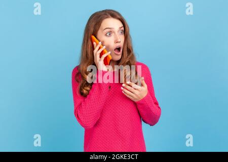 Porträt einer schockierten, wunderten Frau, die einen rosa Pullover trägt, ein Telefongespräch führt, Informationen mit überraschendem Ausdruck hört, unglaubliche Nachrichten. Innen Stockfoto