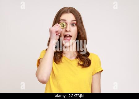 Porträt einer verspielten, aufgeregten jungen Frau mit einem charmanten Lächeln in gelbem T-Shirt, das ein Auge mit goldenem Bitcoin und Kryptowährung bedeckt. Innen Stockfoto