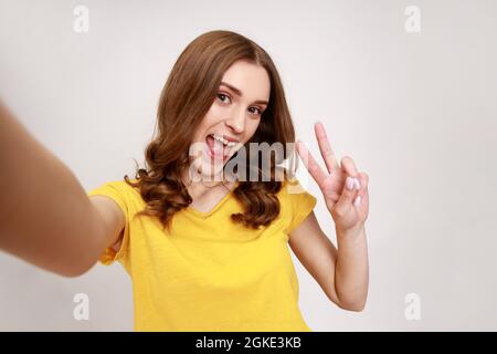 Schöne aufgeregt Teenager-Mädchen in ngelben Casual T-Shirt Aufnahme Selfie, Blick auf Kamera POV, Standpunkt des Fotos, zeigt Sieg Geste. Innen Stockfoto