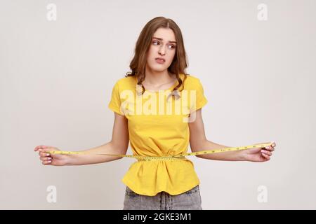 Portrait unglücklich lächelnde Frau in gelbem T-Shirt, die mit einem Maßband ihre Taille misst und Traurigkeit ausdrückt und mit einem Stirnrunzeln wegschaut. Innen Stockfoto