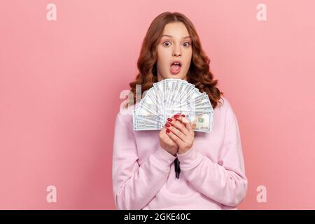 Portrait of stauned Lucky teen girl in Hoodie überrascht, einige Dollar-Scheine in der Hand zu halten, schockiert durch große Lotterie gewinnen, finanzielle Einsparungen. Indoor st Stockfoto