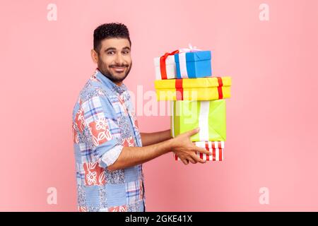 Seitenansicht eines bärtigen Mannes, der einen Stapel Geschenke in den Händen hält, mit einem angenehmen Lächeln auf die Kamera blickt, den Urlaub feiert und ein blaues legeres Hemd trägt. Zoll Stockfoto