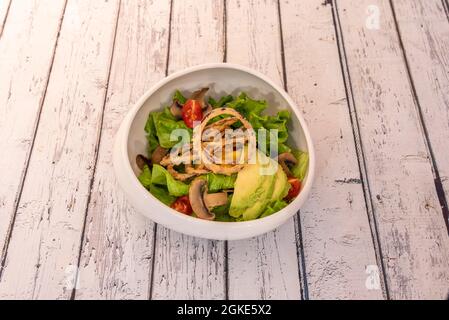 Avocado- und Mangosalat mit zerschlackten Zwiebelringen, Eisbergsalat, in Scheiben geschnittenen Pilzen und gehackten Kirschtomaten in einer weißen Schüssel. Stockfoto