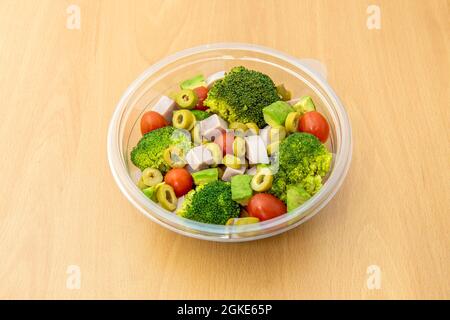 Brokkoli-Salat mit Schinken, gehackten grünen Oliven, reifen Avocados, Kirschtomaten mit Salz und Öl in einem transparenten Kunststoffbehälter für die Lieferung nach Hause Stockfoto