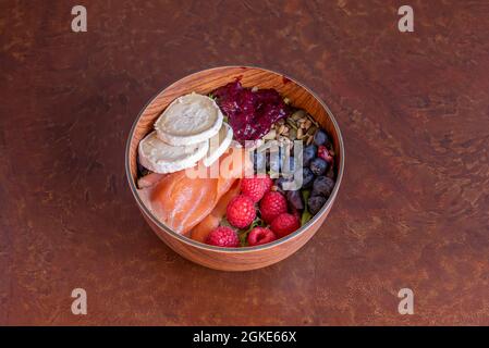 Holzschale mit geräuchertem Lachssalat, Ziegenkäse-Scheiben, Beeren, Sonnenblumen- und Kürbiskernen, Salat und Erdbeermarmelade Stockfoto