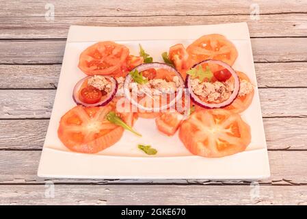 Salat aus geschnittenen geschälten Tomaten, Kirschtomaten, roten Zwiebelringen und Thunfischkonserven auf weißem quadratischem Teller Stockfoto