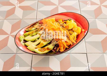 Mangosalat, reife Avocados, Sonnenblumenkerne, geriebene Karotten und Salat mit Balsamico-Essig auf einer länglichen roten Schüssel und Steingut-Tisch Stockfoto