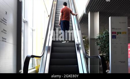 Mann in einem roten T-Shirt, der die Rolltreppe im Business Center hochfährt. HDR. Junger Mann auf der Rolltreppe im Kaufhaus Stockfoto