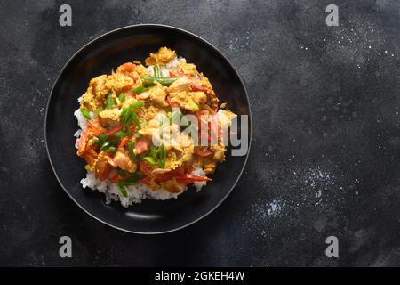 Thailändisches Frühstück Gai Pad Pongali mit Huhn, Eiern, würziger, gelber thai-Paprikapaste, Tomaten, Reis in schwarzer Schüssel im Wok. Nahaufnahme. Panasiatische Küche. Stockfoto