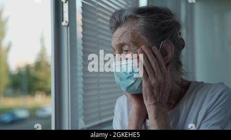Besorgte ältere Frau in einer schützenden medizinischen Maske schaut traurigerweise aus dem Fenster mit ihrem Kopf in den Händen in einem Pflegeheim. Depressive Dame bei Stockfoto