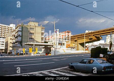 Ein örtlicher Einschienenzug auf der Brücke über einer Straße, Tokio, Japan ca. 1982. Die Monorail in Tokio wurde 1964 eröffnet (um die Olympischen Spiele in diesem Jahr zu nutzen). Es verbindet Hamamatsucho mit dem Flughafen Hameda. Die Züge wurden von Hitachi nach deutschem ALWEG-Design gebaut. Es gilt als die verkehrsreichste (und profitabelste) Monorail der Welt. Eine Tankstelle befindet sich auf der Straße unten. Dieses Bild stammt von der Agfa-Farbtransparenz eines Amateurfotografen – einem Vintage-Foto aus den 1980er Jahren. Stockfoto