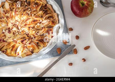 Apfel- und Quark-Tarte mit Beeren und Nüssen in Backform, einige Teller, Messer, Löffel und Apfel auf einem weißen Tisch. Stockfoto