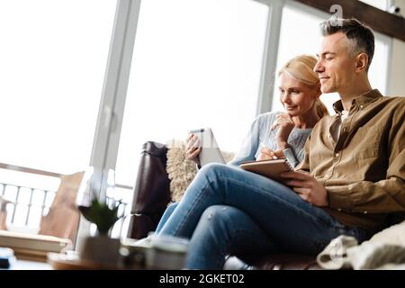 Lächelndes weißes Paar mittleren Alters, das sich zu Hause auf einer Couch entspannt, sich entspannt, mit einem Tablet-PC unterwegs ist und in einem Notebook schreibt Stockfoto