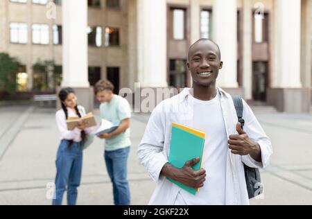Glücklicher afroamerikanischer Student, der draußen mit Klassenkameraden auf dem Universitätscampus steht, posiert und vor der Kamera lächelt Stockfoto