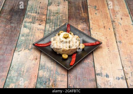 Typisch spanische Tapa aus russischem Salat mit gekochtem Ei, Oliven, roten Paprika und gekochten Kartoffeln mit Mayonnaise auf einem quadratischen schwarzen Teller Stockfoto