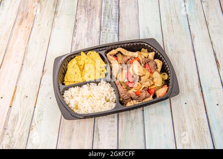 Schwarzes Liefertablett mit gemischten Fajitas aus Huhn und Rind mit Mais-Nachos und weißem Reis auf Holzhintergrund Stockfoto