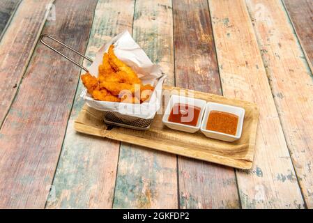 Frittierkorb aus Metall, gefüllt mit Hähnchenfingern mit zwei Dipping-Saucen auf einem Holztisch Stockfoto