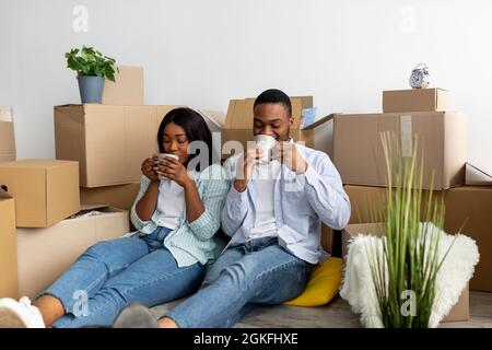 Entspanntes schwarzes Paar, das zwischen Kartons sitzt und Kaffee trinkt, nachdem es in eine neue Wohnung umzog Stockfoto