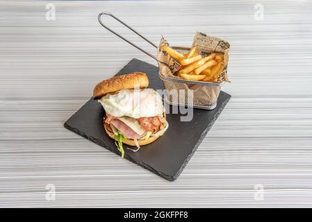 Köstlicher Rindfleisch-Burger mit Spiegelei, so dass sie versuchen, es zu essen, ohne es zu färben, begleitet von einem Korb mit pommes auf dem grauen Tisch Stockfoto