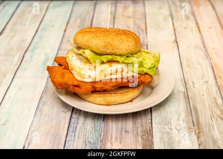 Einsamer Hamburger mit Hähnchenfilet im Teig mit einem ganzen Spiegelei mit Salat und Brot mit Sesam auf Holztisch Stockfoto