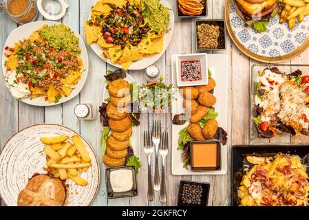 Abwechslungsreiche Gerichte und Hamburger aus der Sicht von oben, mit Kroketten, Guacamole und Nachos, Kartoffeln mit Speck und Käse, Arapas und einem Holzhintergrund Stockfoto