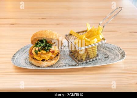 Spektakulärer Rindfleischburger mit viel Käse, gebratener Rucola und italienischer dehydrierter Tomate mit einer Portion Pommes als Beilage auf einem Holztisch Stockfoto