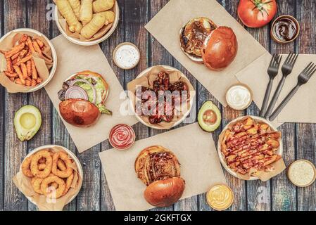 Draufsicht auf Fast-Food-Gerichte, bbq Chicken Wings, verschiedene Rindfleisch-Burger, Zwiebelringe, tequeños, Kartoffeln mit Soßen und Avocado Stockfoto