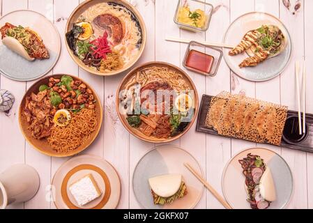 Teller mit typisch koreanischen und japanischen Speisen mit vielen Ramen, Nudeln, Gyozas, bao-Brot-Sandwiches, Krabben und Radieschen auf weißem Tisch Stockfoto