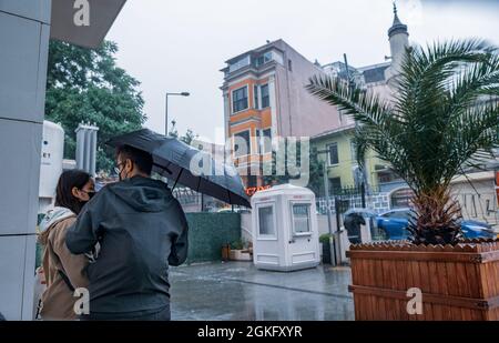 Beyoglu, Istanbul, Türkei - 06.18.2021: Männer- und Frauenfreunde mit schwarzem Regenschirm warten in der Nähe eines Gebäudes, um sich vor heftigem Regen draußen in Taken zu schützen Stockfoto